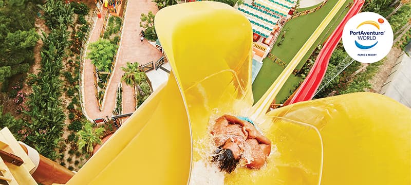 Aerial view of a bright yellow water slide at PortAventura World water park, with a rider splashing into the pool below and lush green surroundings nearby. The PortAventura World logo is visible in the top right corner.